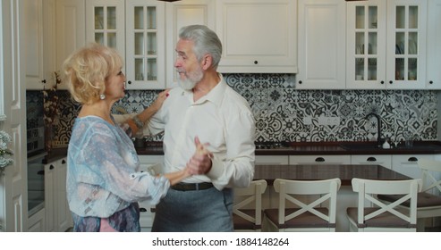 Mature Senior Couple Dancing In The Kitchen, Beautiful Romantic Older Grandparents Relaxing Having Fun Together At Home Celebrating Anniversary Enjoy Care Love Tenderness.