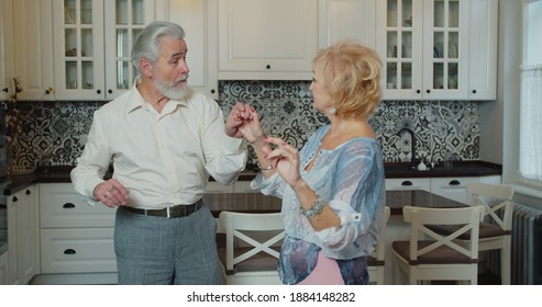 Mature Senior Couple Dancing In The Kitchen, Beautiful Romantic Older Grandparents Relaxing Having Fun Together At Home Celebrating Anniversary Enjoy Care Love Tenderness.