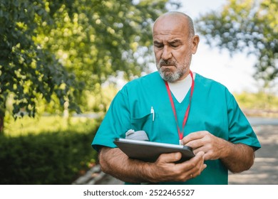 Mature senior man doctor in green medical scrubs stand outdoor with a focused expression, examining a tablet, drink coffee while have break from work - Powered by Shutterstock