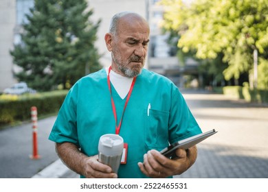 Mature senior man doctor in green medical scrubs stand outdoor with a focused expression, examining a tablet, drink coffee while have break from work - Powered by Shutterstock