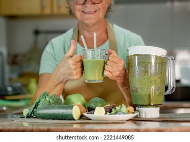 Mature senior woman drinking healthy green smoothie holding a glass with straws. Smiling caucasian lady in home kitchen drinking detox juice