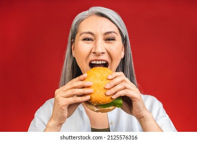 Mature Senior Woman Eating Burger With Satisfaction. Grandmother Enjoys Tasty Hamburger Takeaway, Delicious Bite Of Burger, Order Fastfood Delivery While Hungry, Standing Isolated Over Red Background.