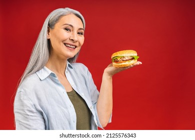 Mature Senior Woman Eating Burger With Satisfaction. Grandmother Enjoys Tasty Hamburger Takeaway, Delicious Bite Of Burger, Order Fastfood Delivery While Hungry, Standing Isolated Over Red Background.