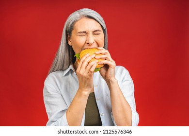 Mature Senior Woman Eating Burger With Satisfaction. Grandmother Enjoys Tasty Hamburger Takeaway, Delicious Bite Of Burger, Order Fastfood Delivery While Hungry, Standing Isolated Over Red Background.