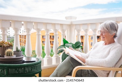 Mature senior woman relaxing on home balcony reading a book after work - attractive gray haired adult female people enjoying peaceful lifestyle outdoors