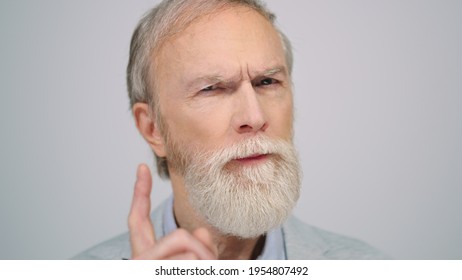 Mature serious man looking camera in studio grey background. Focused old gentleman wagging finger indoors. Disappointed male person frowning to camera inside.