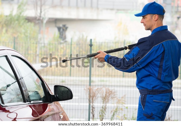 Mature serviceman with high pressure water jet\
washing red car at service\
station