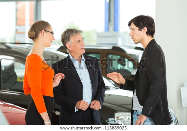 Mature single man with autos in light car\
dealership with a young couple,- man, young -, he obviously is\
buying a car or is a car\
dealer