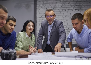 Mature skilled proud ceo in eyeglasses with team of male and female experienced professionals cooperating on architectural sitting at desktop with sketch of blueprint in stylish office interior - Shutterstock ID 697584031