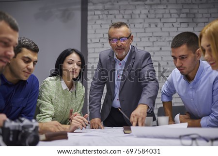 Mature skilled proud ceo in eyeglasses with team of male and female experienced professionals cooperating on architectural sitting at desktop with sketch of blueprint in stylish office interior