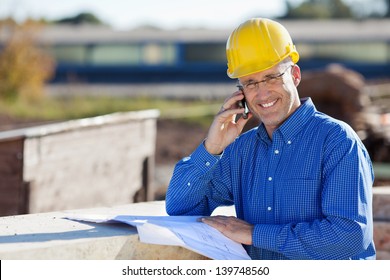 Mature smiling architect while using mobile phone at construction site