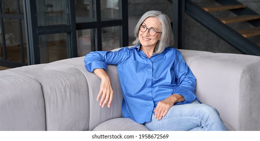 Mature Smiling Peaceful 60s Middle Age Lady Sitting On Couch Relaxing At Home. Senior Elder Serene Woman In Glasses Looking Away Thinking Of Positive Vision, Dreaming Of Future And Enjoying Wellbeing.