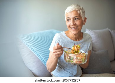 Mature Smiling Woman Eating Salad, Fruits And Vegetables. Attractive Mature Woman With Fresh Green Salad At Home. Senior Woman Relaxing At Home While Eating A Small Green Salad, Home Interior.