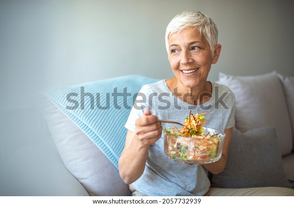 Mature smiling woman eating salad, fruits and
vegetables. Attractive mature woman with fresh green salad at home.
Senior woman relaxing at home while eating a small green salad,
home interior.