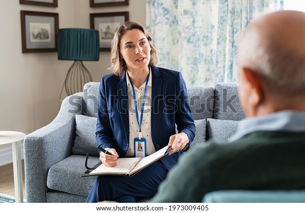 Mature social worker talking to senior man and
taking notes of health progress. Mid adult woman in formal clothing
visiting old man at home for medical history. Support worker
talking to elder.