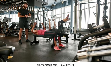 Mature Sporty Couple Lifting Dumbbels Lying On Workout Bench In Large Gym. Young Confident Trainer Is Timing, Using Hand Watch. Setting Personal Goals For Clients.