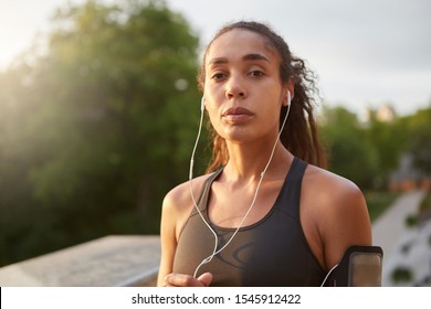 Mature sporty model with casual hairstyle going in for sports in green city park, wearing black sporty top and earphones, looking at camera with folded lips