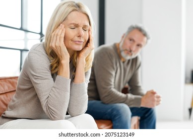 Mature spouses close to divorce, frustrated wife sits with eyes closed, upset grey-haired husband on the background. Middle-aged couple sits separate on the sofa at home. Misunderstanding each other