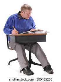 A Mature Student Hard At Work As He Sits In A Retro Child's School Desk,  On A White Background.