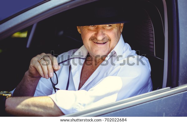 Mature stylish European or\
American man wear black hat, glasses and white shirt driven car,\
modern and hipster style for older people. Such a good mood at\
pretty day