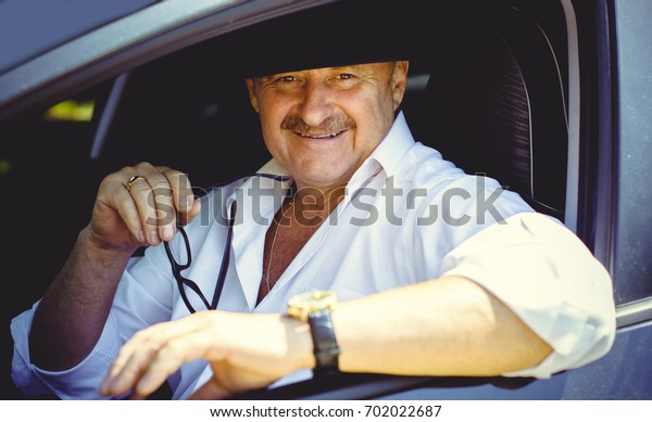 Mature stylish European or\
American man wear black hat, glasses and white shirt driven car,\
modern and hipster style for older people. Such a good mood at\
pretty day