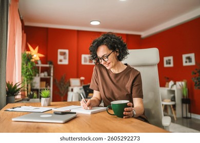 Mature stylish woman take notes in notebook at home. Female freelancer write details on notebook while work from home in living room.