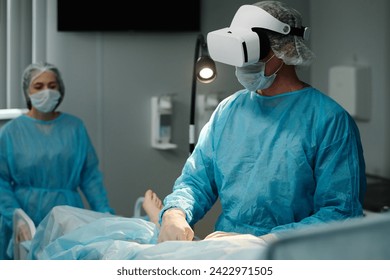 Mature surgeon in VR headset, scrubs, protective mask and gloves using medical equipment while performing surgical operation - Powered by Shutterstock