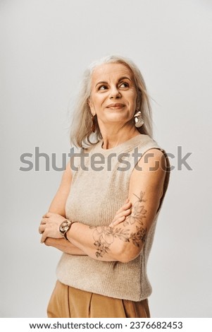 mature tattooed lady in knitted top standing with folded arms and looking away on grey, positivity