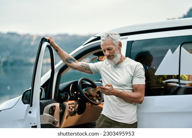 Mature tattooed man standing near rv camper van on vacation using mobile phone. Smiling mature active traveler holding smartphone enjoying free internet in camping tourism nature park. - Powered by Shutterstock