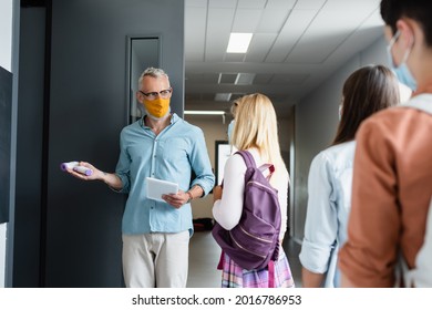 Mature Teacher With Pyrometer And Digital Tablet Inviting Schoolgirl In Medical Mask Into Classroom