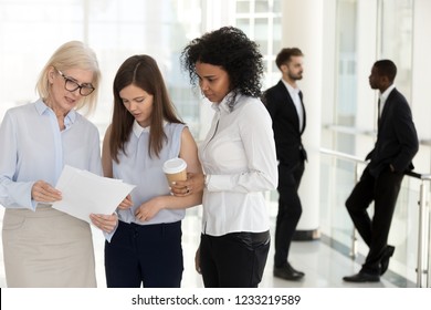 Mature team leader and young female employees discussing paperwork standing in office, diverse employees talking about document, executive giving instructions explaining work to colleagues interns