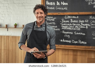 Mature waiter wearing black apron and standing in front of the blackboard with the menu of the day. Portrait of smiling man holding digital tablet and looking at camera. Happy small business owner. - Powered by Shutterstock