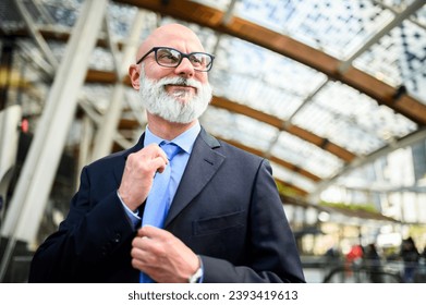 Mature, well-dressed businessman adjusts his tie in a modern glass-roofed atrium - Powered by Shutterstock