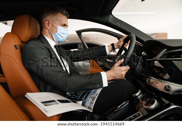 Mature white man in face mask choosing and\
examining car at\
showroom
