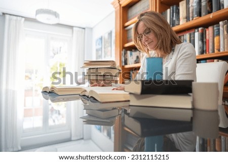 Mature white woman with several books on a glass table and daylight pouring in through the window.