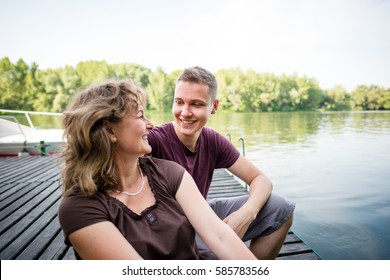 Mature Woman With Adult Son Sitting On Wooden Jetty, Tranquil Surrounding.