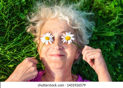 Mature Woman After 60 Is Lying On The Lawn Holding Chamomile Eyes. Care For Women's Wrinkled Skin. Vision And Eye Care. Women Health