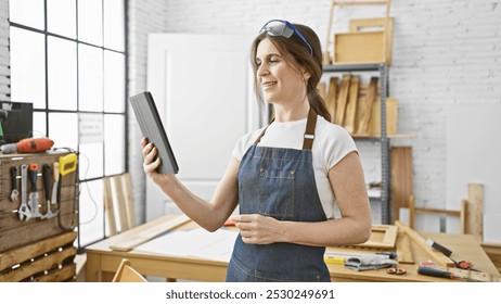 Mature woman in apron holding tablet in woodshop with tools, standing confidently in her creative workshop. - Powered by Shutterstock