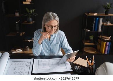 Mature woman, architect and reading with tablet, screen and ideas with review, floor plan or property in office. Person, ux and blueprint with design, drawing or development for real estate project