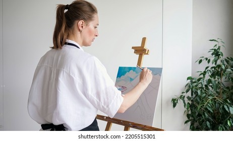 Mature Woman Artist With Ponytail Paints Sky Landscape On Canvas. Skilled Lady Master In White Enjoys Painting Picture On Wooden Easel Closeup