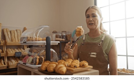 Mature woman in a bakery indoor selecting fresh pastries with tongs, surrounded by bread and various baked goods on display. - Powered by Shutterstock