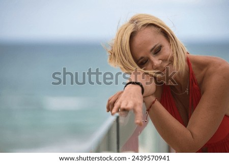 Mature woman, beautiful, blonde, with a red dress, leaning on a glass railing with her eyes closed, relaxed and calm with the sea in the background. Concept beauty, fashion, trend, travel, maturity.
