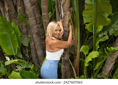 Mature woman, beautiful, blonde, with white top and jeans, in a smiling and funny attitude, clinging to the trunk of a palm tree. Concept beauty, fashion, trend, travel, maturity.