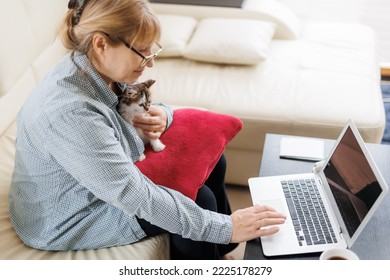 Mature Woman In Blue Shirt Sitting With A Cat On Her Lap At The Wooden Table At Home With Laptop And Notebook, Working Or Shopping Online.