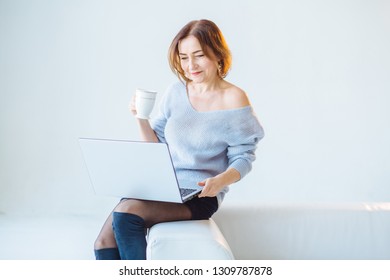 Mature Woman In Blue Sweater Working With Laptop, Drink Coffee Over Light Blue Gray Background. Portrait Of Smiling Happy Shining Senior Business Woman Brows Internet At Home.