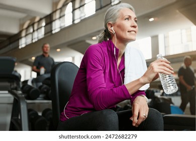 Mature woman, break and drinking with water, earphones or music on machine for workout or exercise at gym. Female person with smile for rest, mineral beverage or thirst after training at health club - Powered by Shutterstock