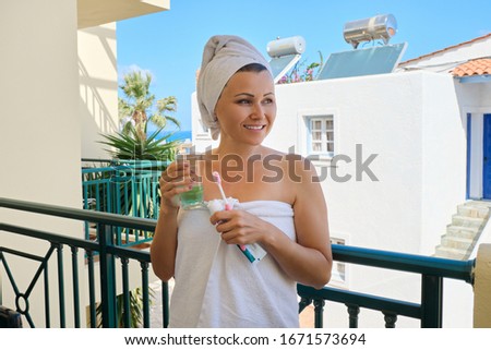 Mature woman brushes her teeth, female with toothbrush glass of water in bath towel on outdoor balcony, sunny summer day at resort