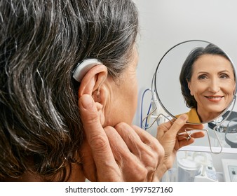 Mature Woman With BTE Hearing Aid Looks At Herself In Mirror And Tries On Hearing Device, Close-up. Hearing Loss Treatment