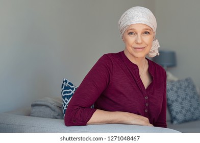 Mature woman with cancer in pink headscarf smiling sitting on couch at home. woman suffering from cancer sitting after chemotherapy sessions. Portrait of mature lady facing side-effects of hair loss.