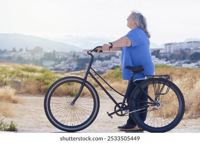 A mature woman carries a black retro-style bicycle, with a view of a small village and mountains with clouds in the background - Powered by Shutterstock